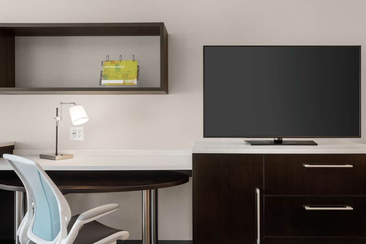 Een moderne kantoorruimte met een bureau, stoel, lamp en een groot flatscreen televisietoestel aan een witte muur.