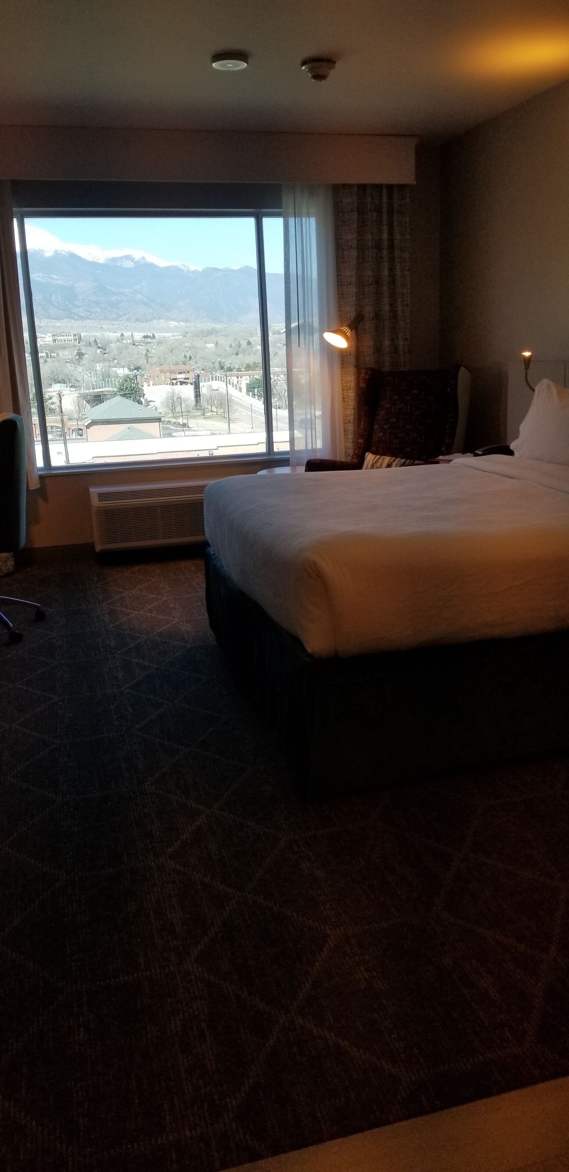 A hotel room with a large bed, a chair, a desk, and a view of mountains through a large window.
