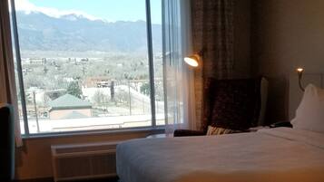 A hotel room with a large bed, a chair, a desk, and a view of mountains through a large window.