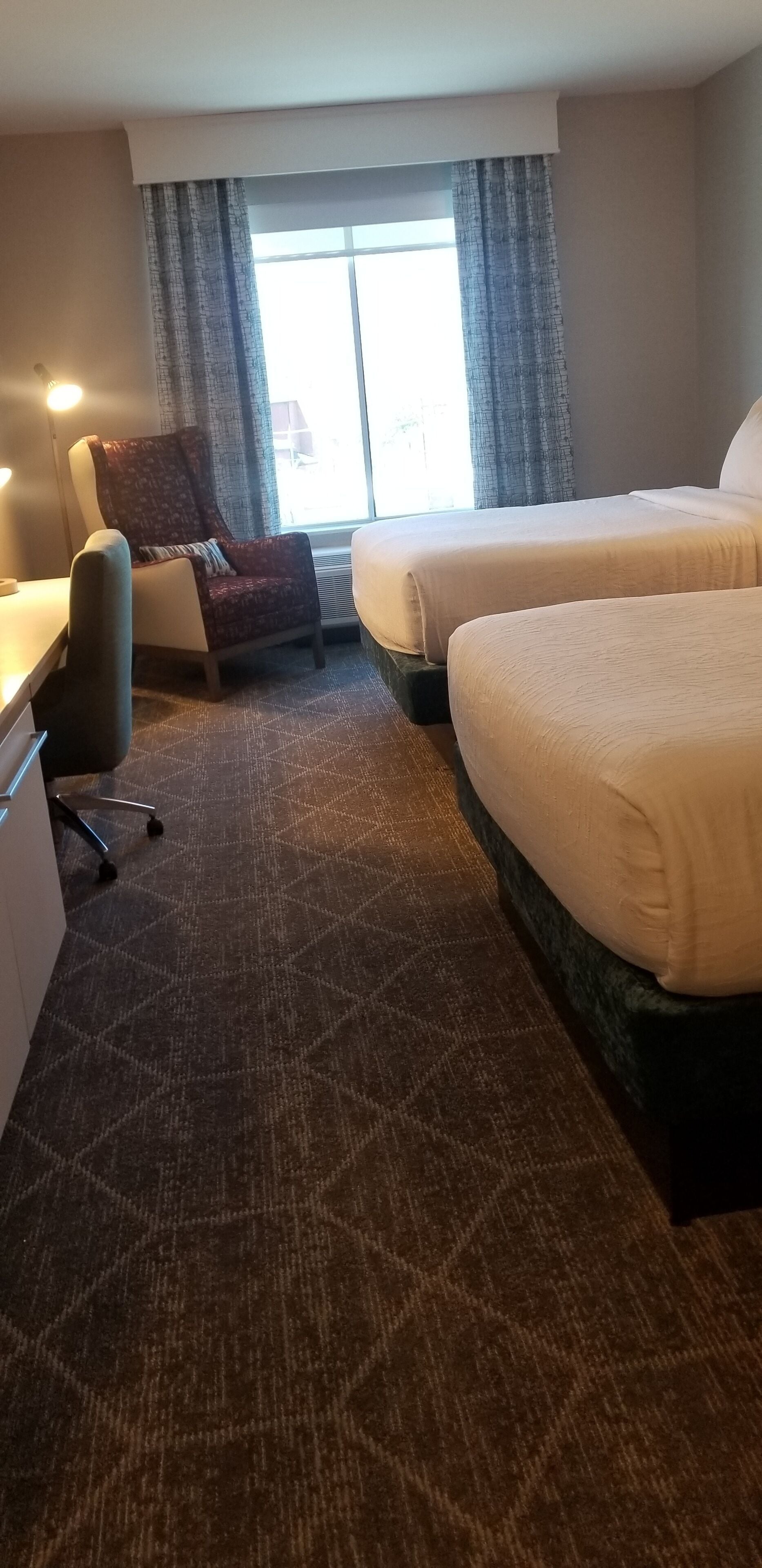 A hotel room with two beds, a desk, a chair, and a large window with curtains.