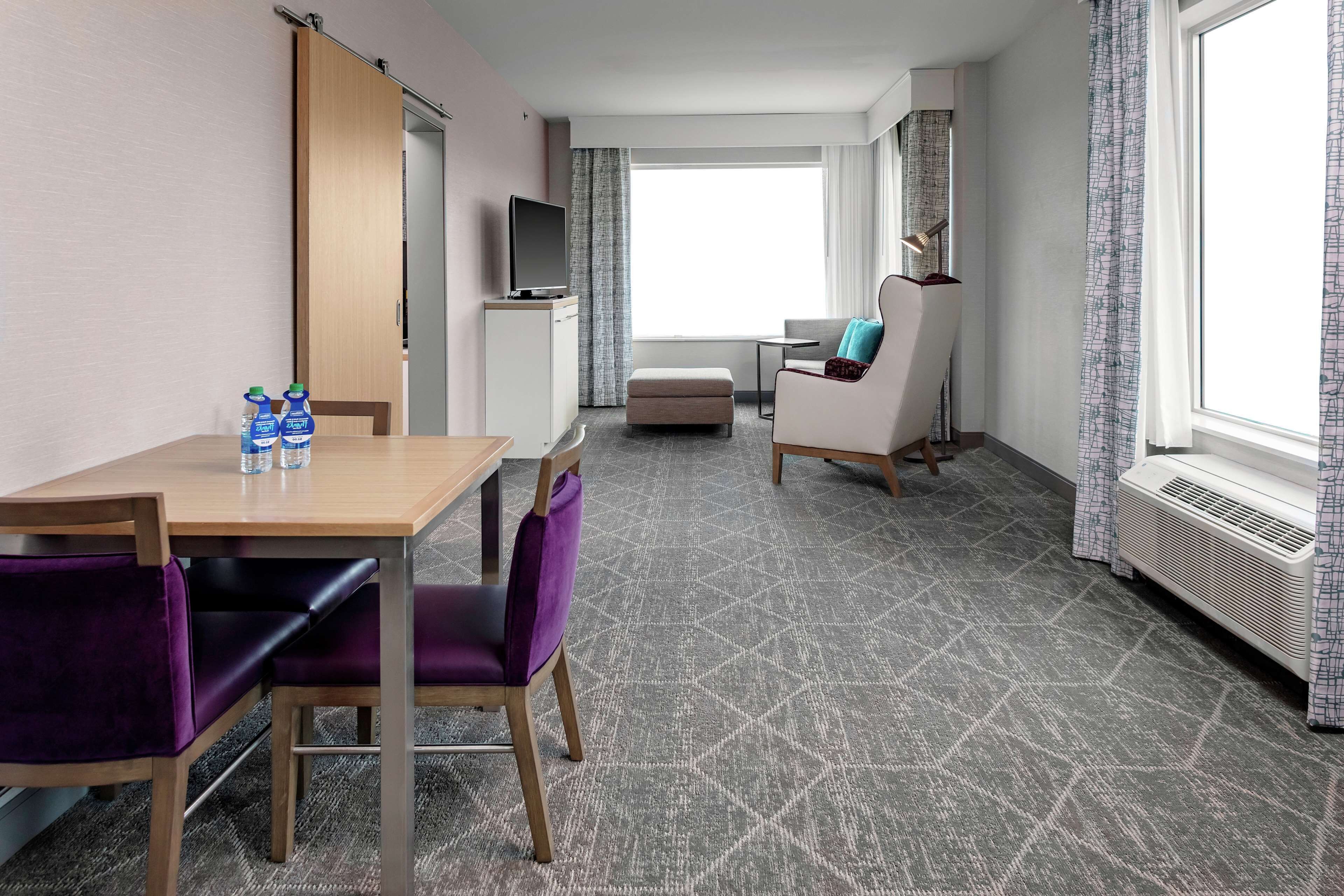 A hotel room with a dining table, purple chairs, a TV, a sofa, and a large window.