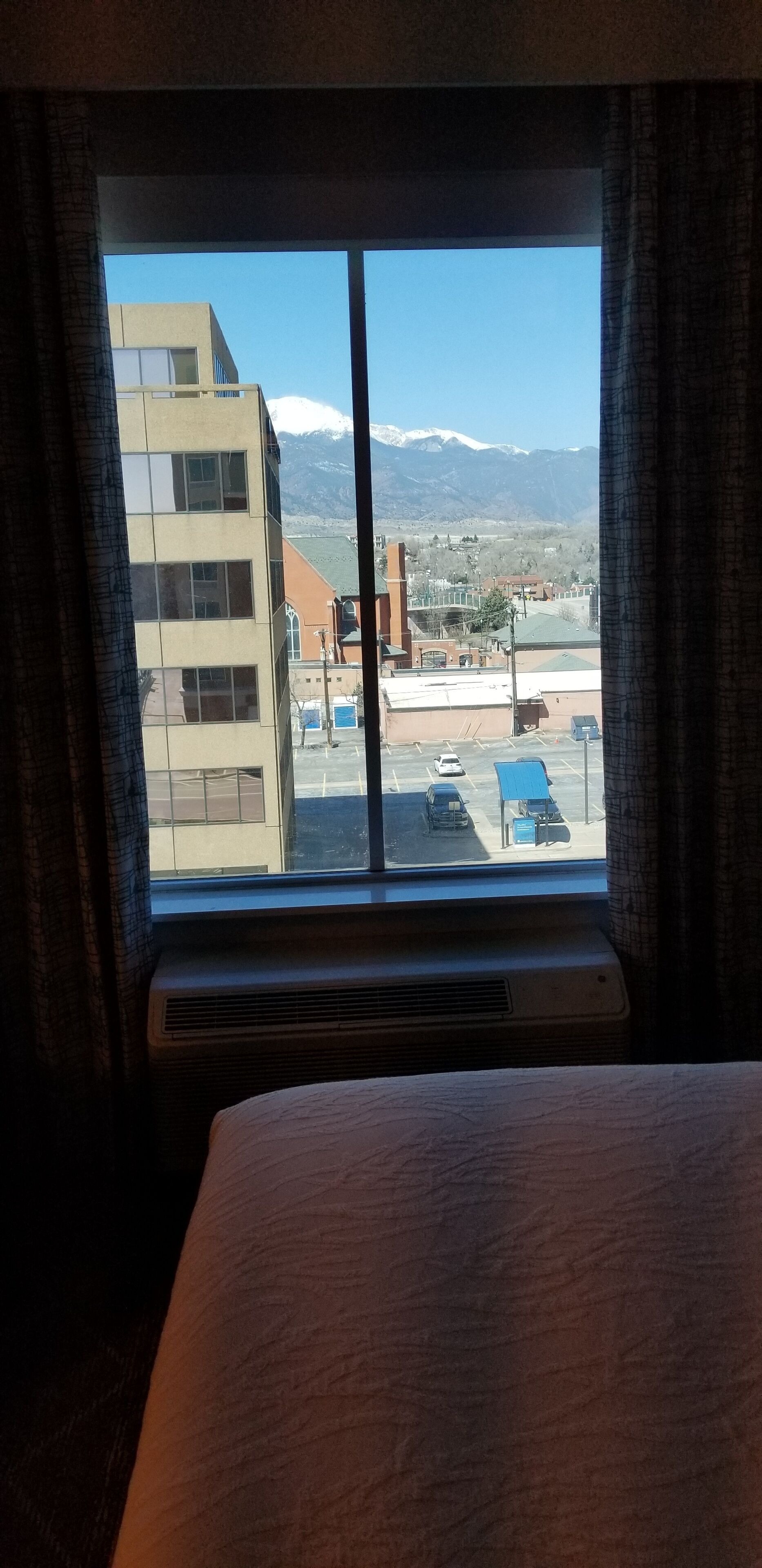 A hotel room with a large window offering a view of a parking lot and buildings, including a mountain range in the distance.