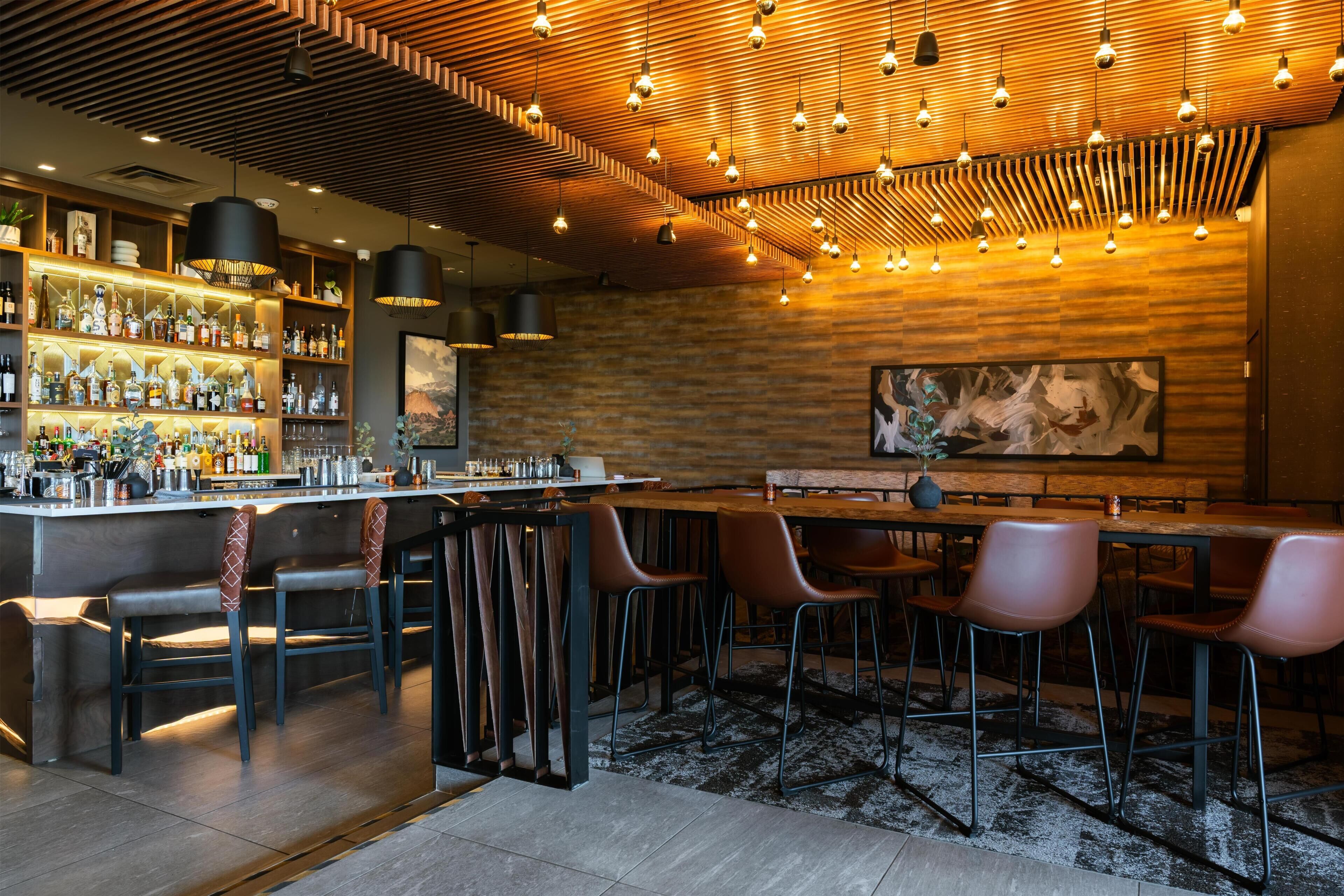 Uma área de bar com teto de madeira, luminárias pendentes e balcão com garrafas e copos.