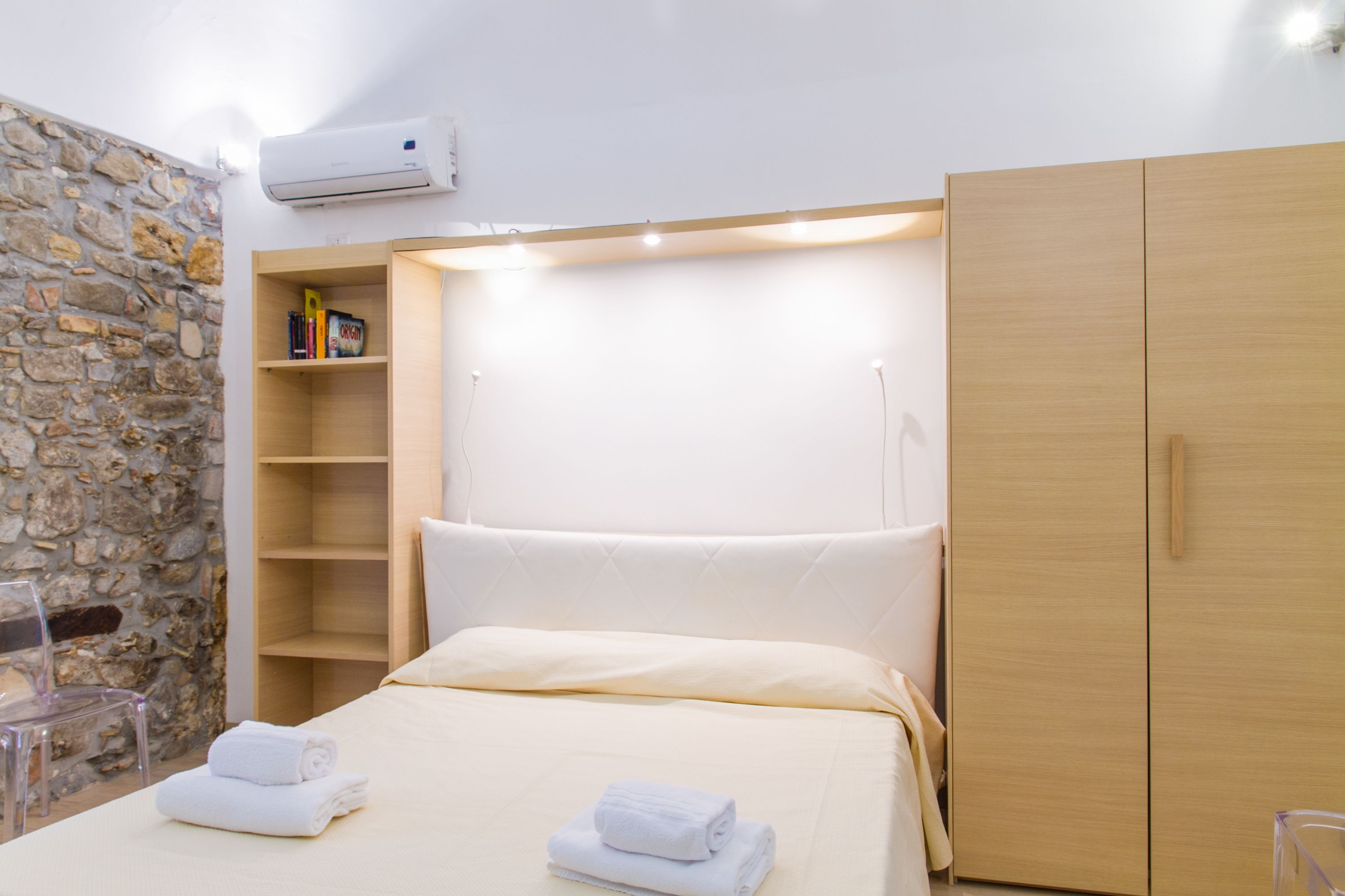 Een hotelkamer met een stenen muur, een houten kledingkast, een bed met witte lakens en een plank met boeken.