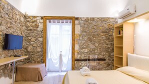 Une pièce avec un mur en pierre, un lit recouvert de draps blancs, un téléviseur posé sur une table en bois et une étagère en bois remplie de livres.