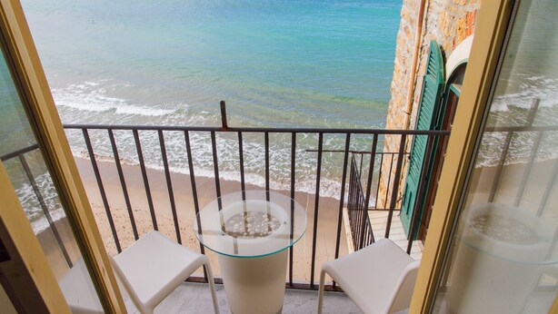 Un balcon avec une table en verre et deux chaises blanches, donnant sur une plage avec des vagues et un bâtiment aux volets verts.
