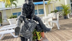 Une statue d’un homme assis sur une terrasse en bois, agrémentée de plantes en pot et de mobilier d’extérieur.