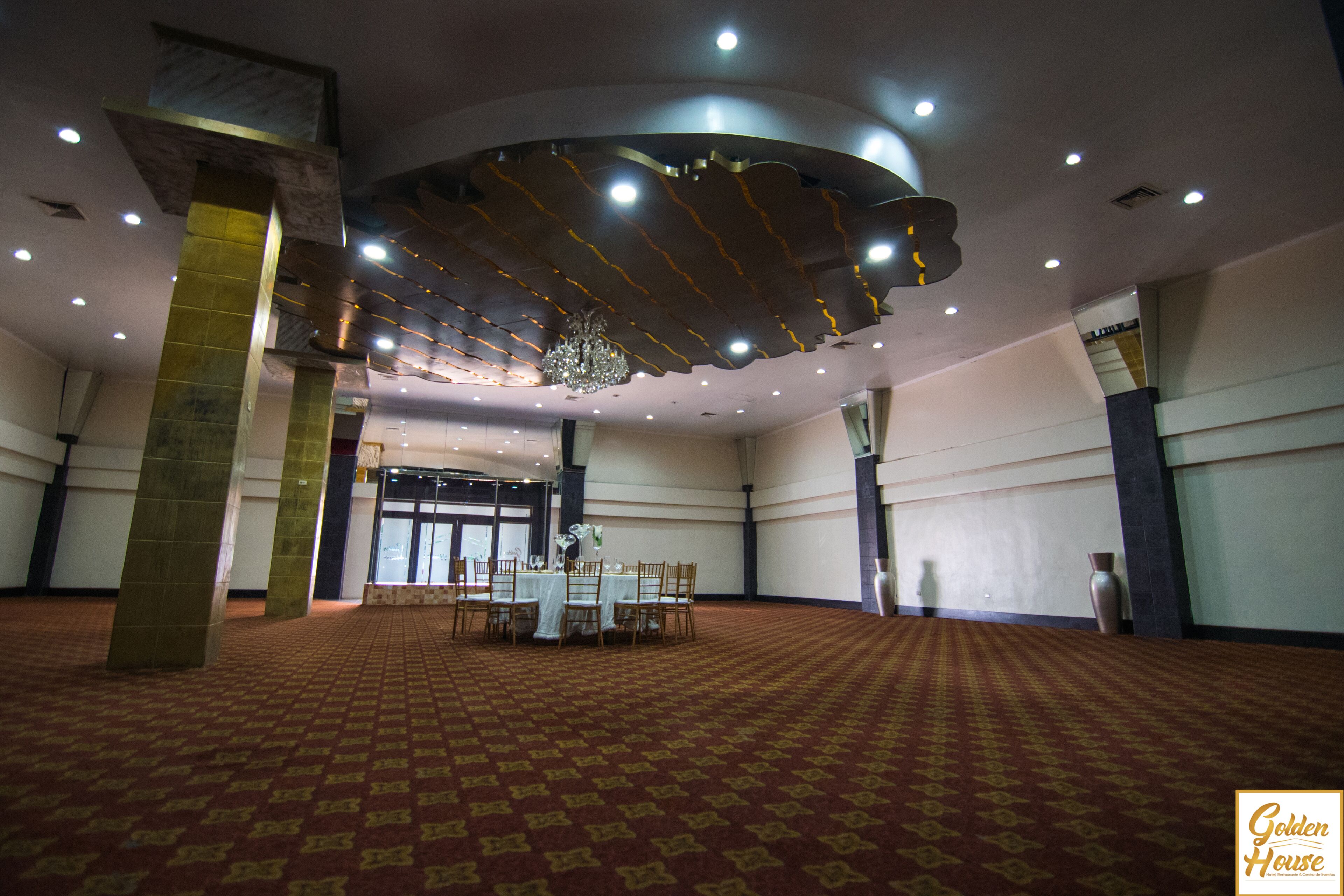 A spacious hall with a patterned carpet, a chandelier, and tables set up for dining.