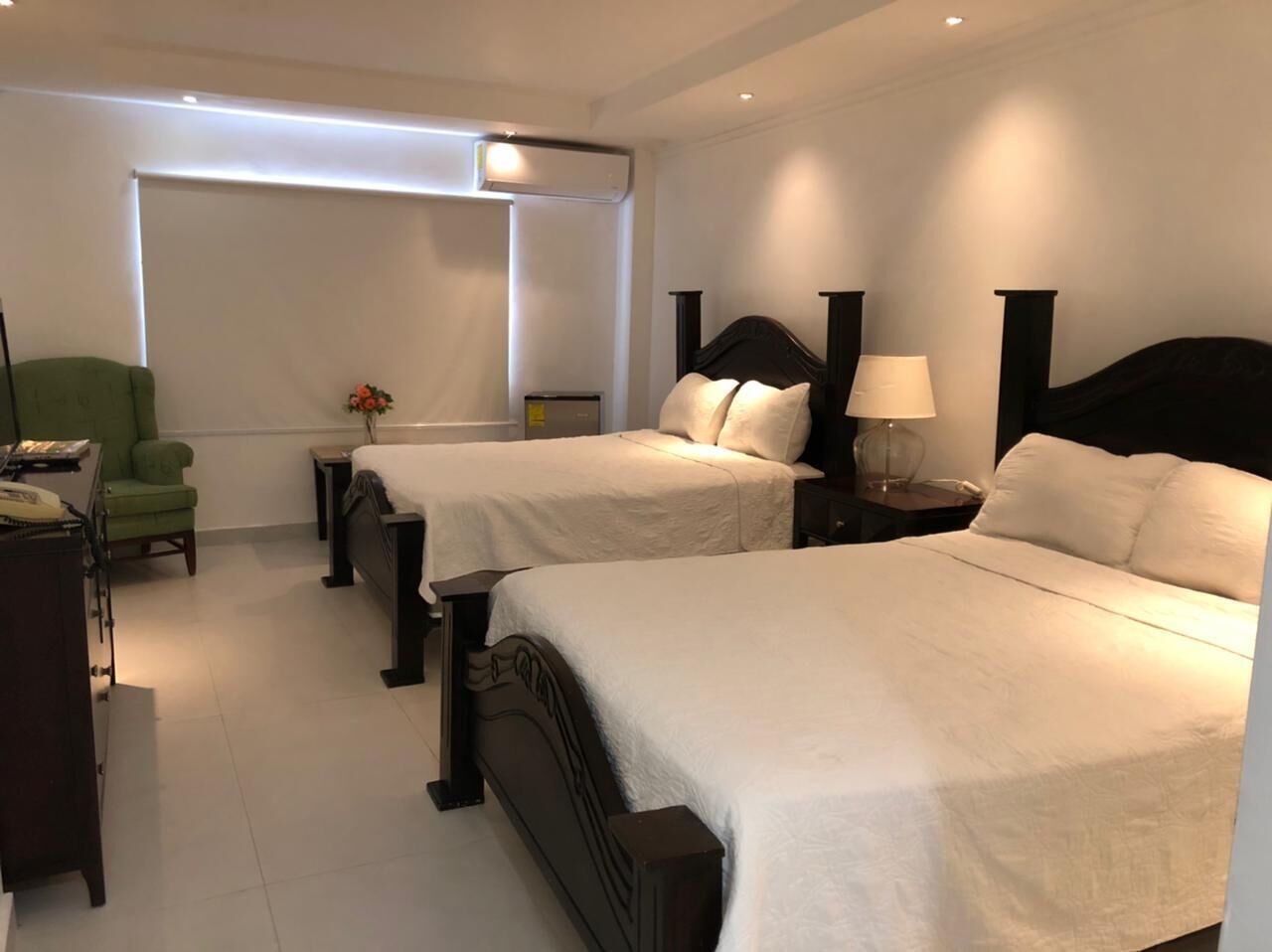 Ein Hotelzimmer mit zwei Einzelbetten, einem Schreibtisch mit Stuhl, einem Nachttisch mit Lampe und einem großen Fenster mit Jalousien.