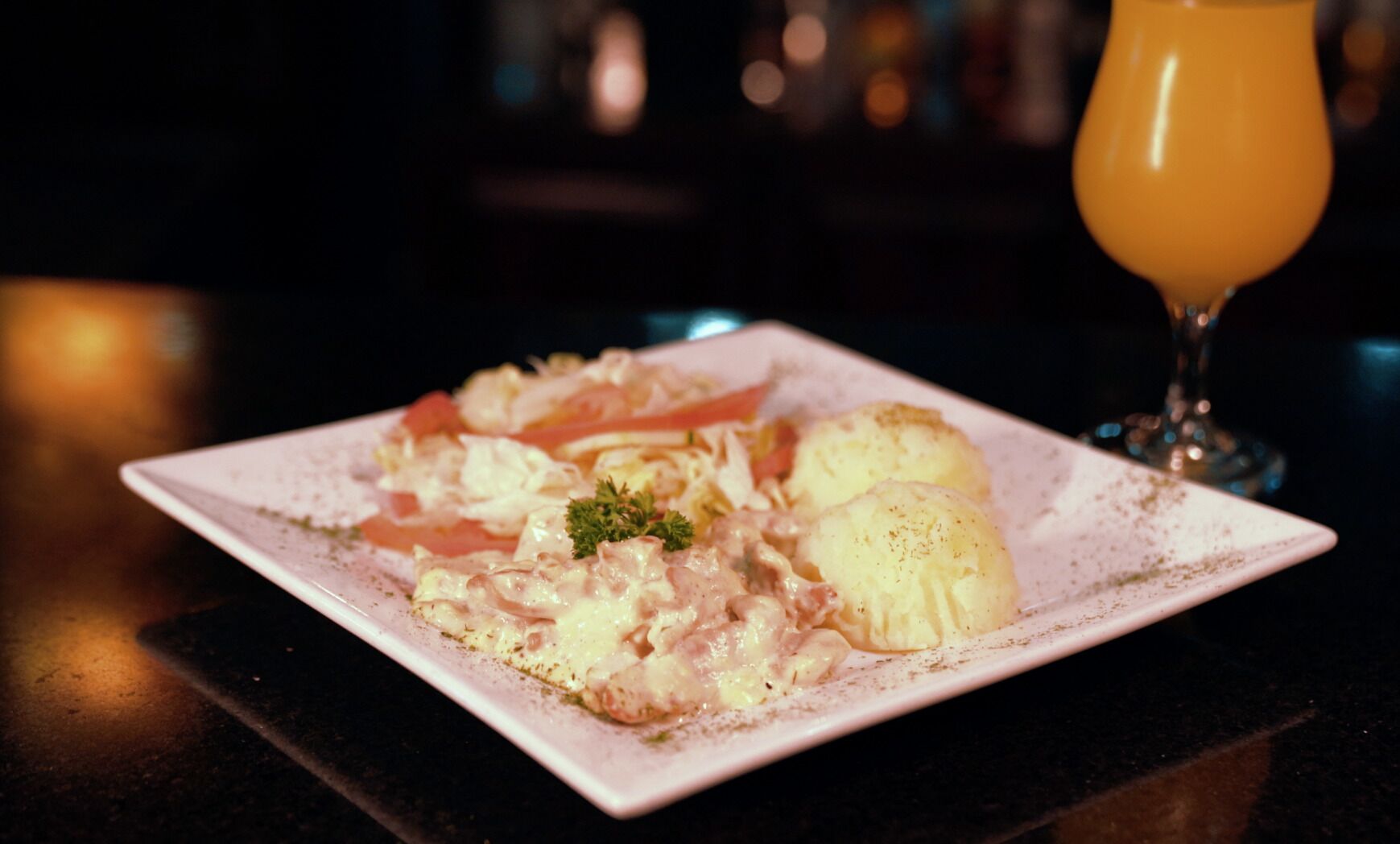 Une assiette carrée contenant un plat crémeux, probablement un gratin, garni de persil, accompagnée de deux accompagnements de pommes de terre en forme de galettes, et un verre de jus d’orange en arrière-plan.