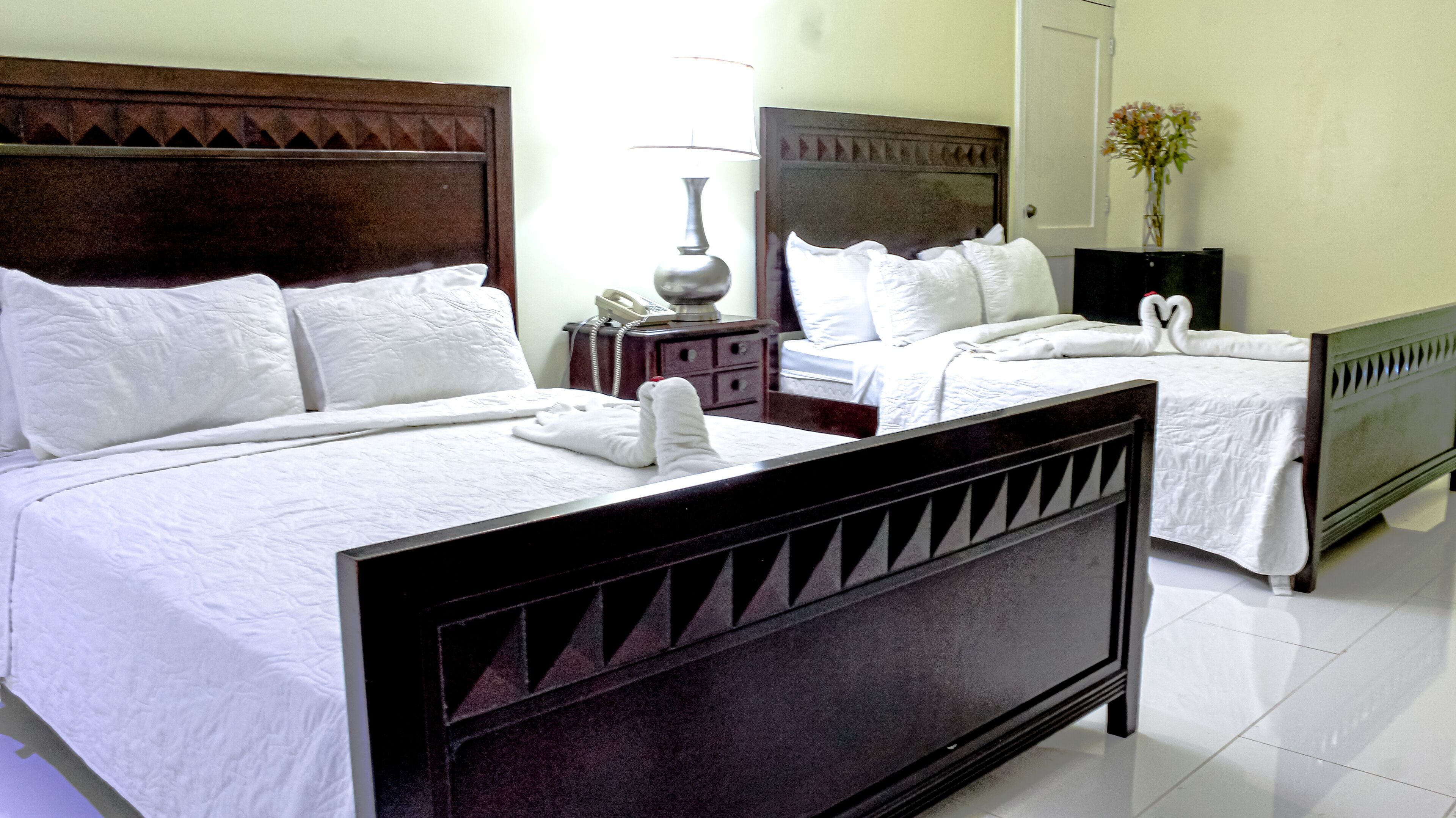 Quarto de hotel com duas camas, cada uma com roupa de cama branca e um travesseiro decorativo em forma de cisne. Há uma mesa de cabeceira com abajur e telefone.