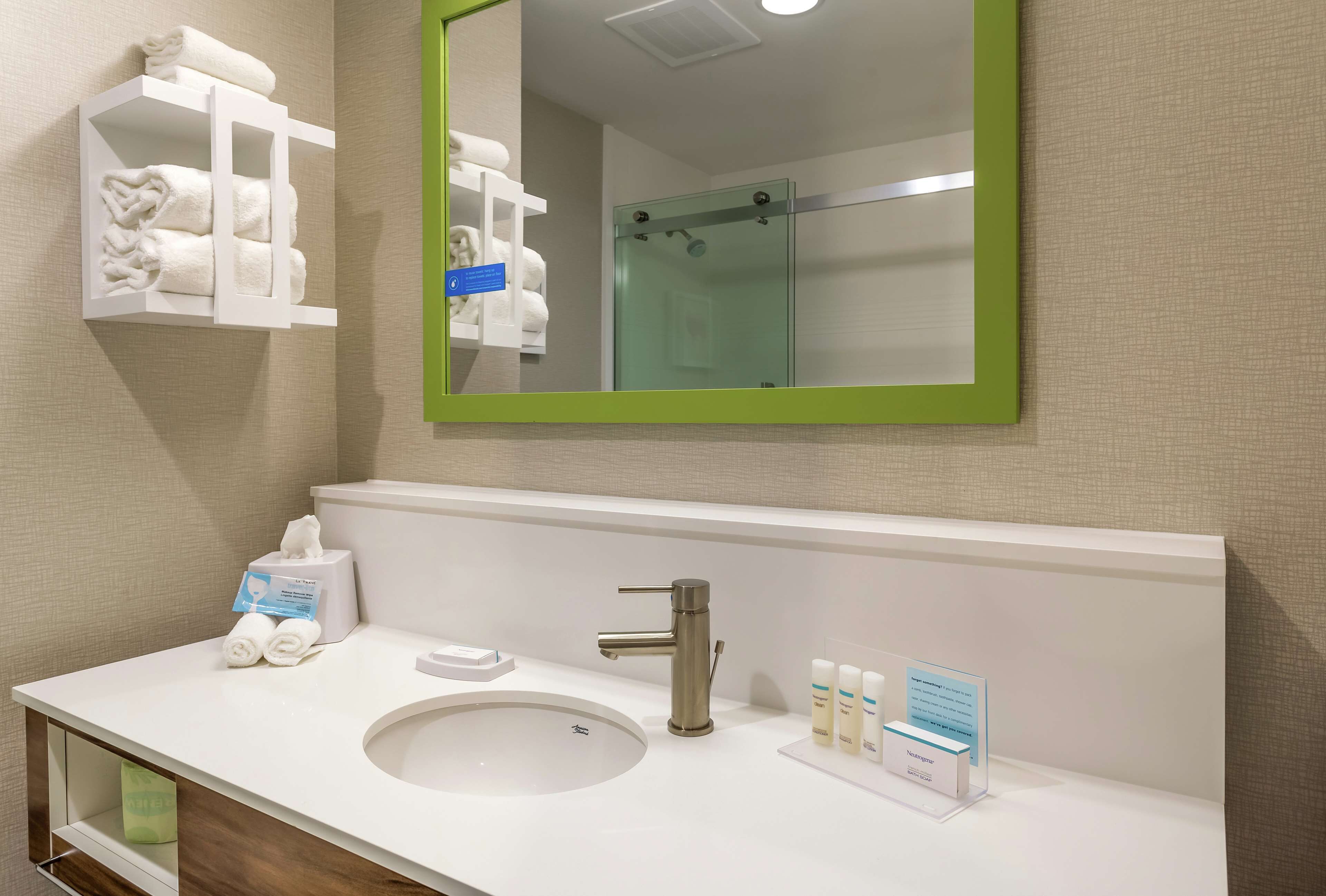 Une salle de bain d’hôtel avec un lavabo blanc, un miroir encadré de vert, une cabine de douche en verre et un porte-serviettes où sont rangées des serviettes pliées avec soin.