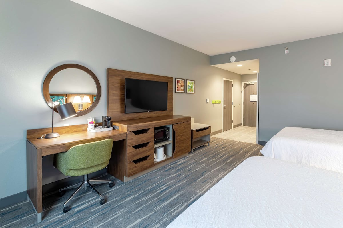 Une chambre d’hôtel avec un bureau en bois, une télévision, un fauteuil vert, un lit et une salle de bain visible au fond.