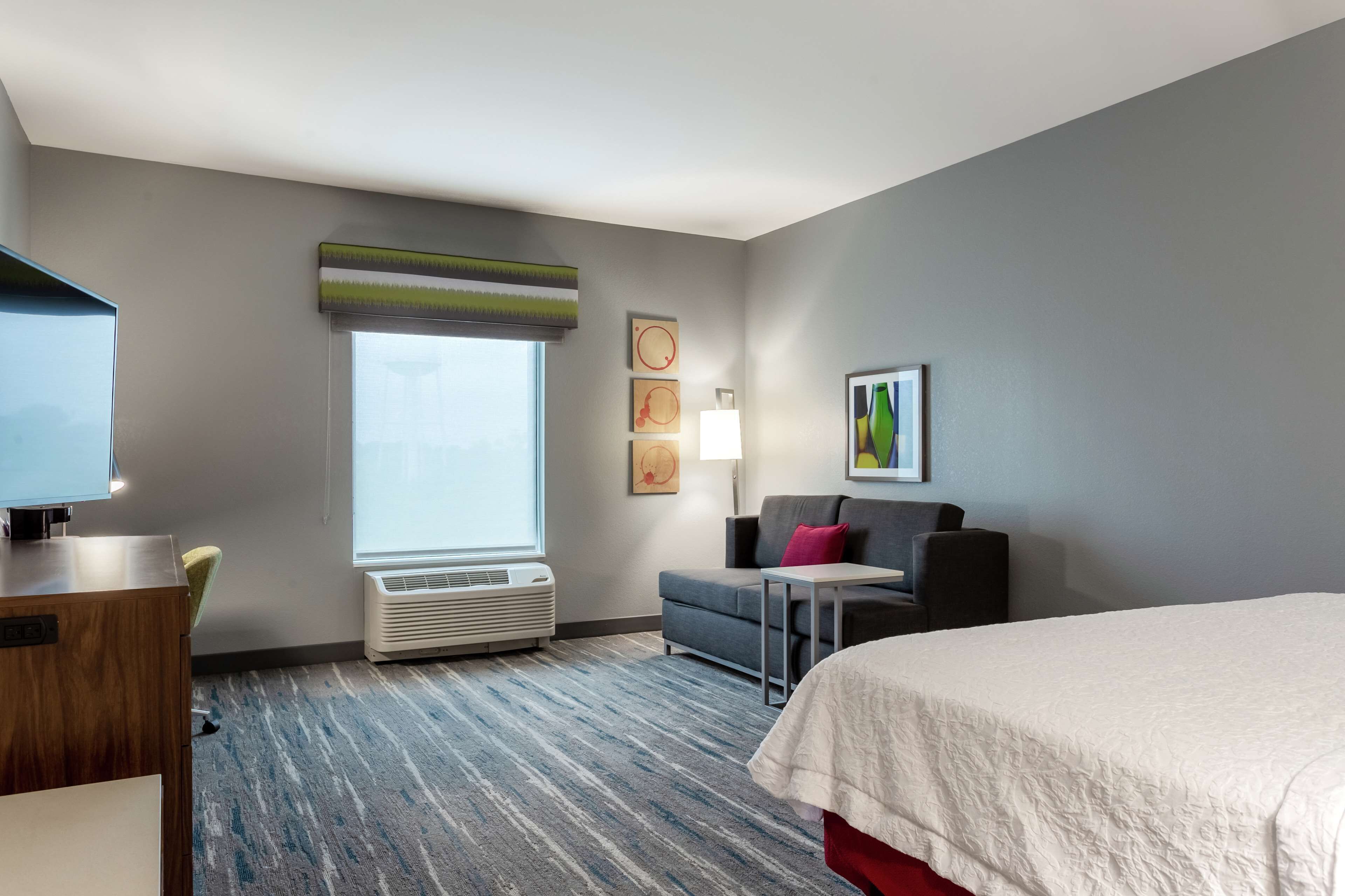 A hotel room with a bed, a desk, a chair, a sofa, a small table, a lamp, and a window with blinds.