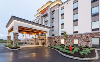 Entrada de un hotel Hampton Inn con un vestíbulo cubierto, fachada de piedra y un hidrante en el jardín.