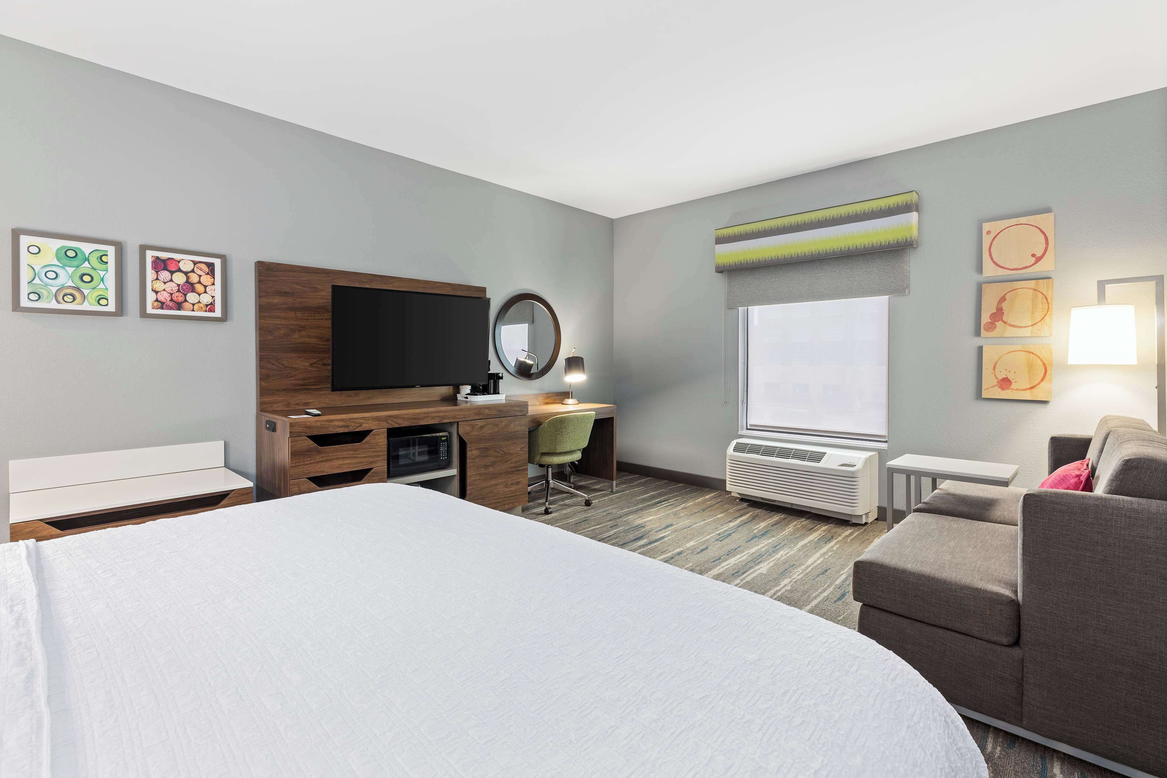 A hotel room with a bed, a desk with a chair, a TV, a mirror, a window, and artwork on the walls.