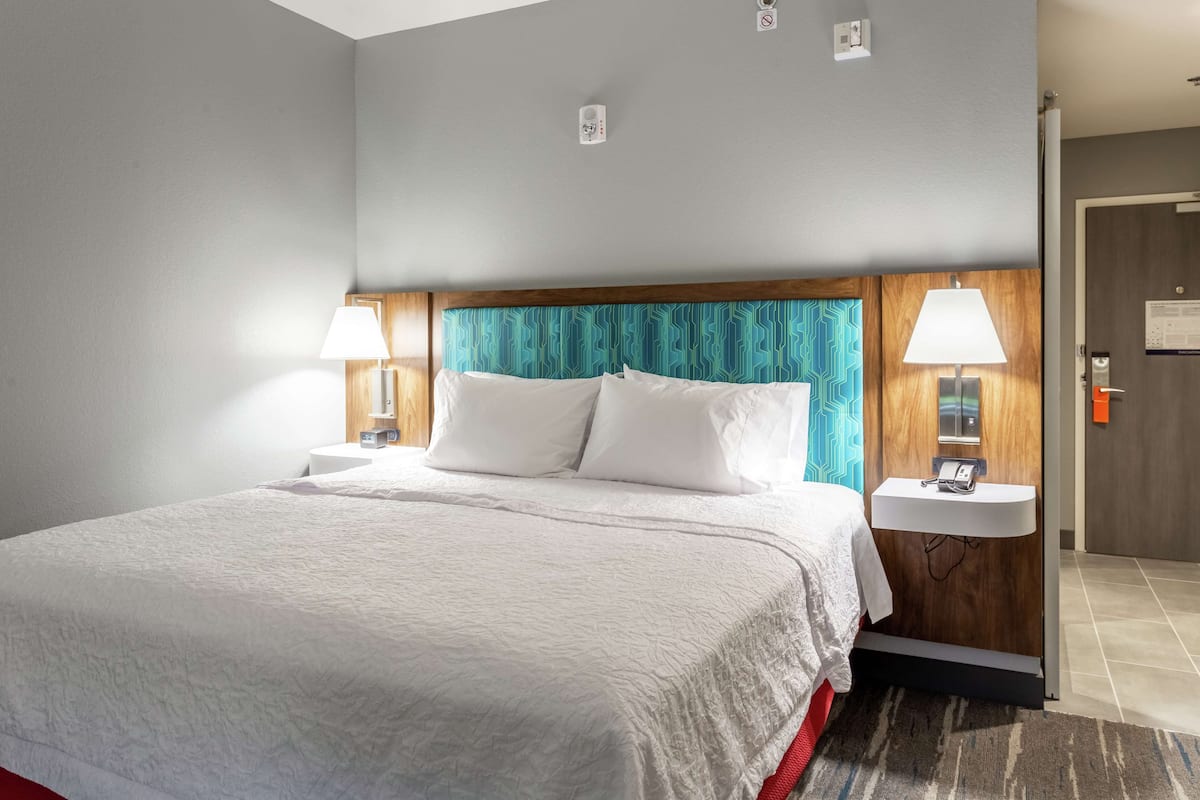 Une chambre d’hôtel avec un grand lit, deux tables de chevet avec des lampes, une tête de lit en bois et une petite salle de bain comprenant un lavabo et un miroir.