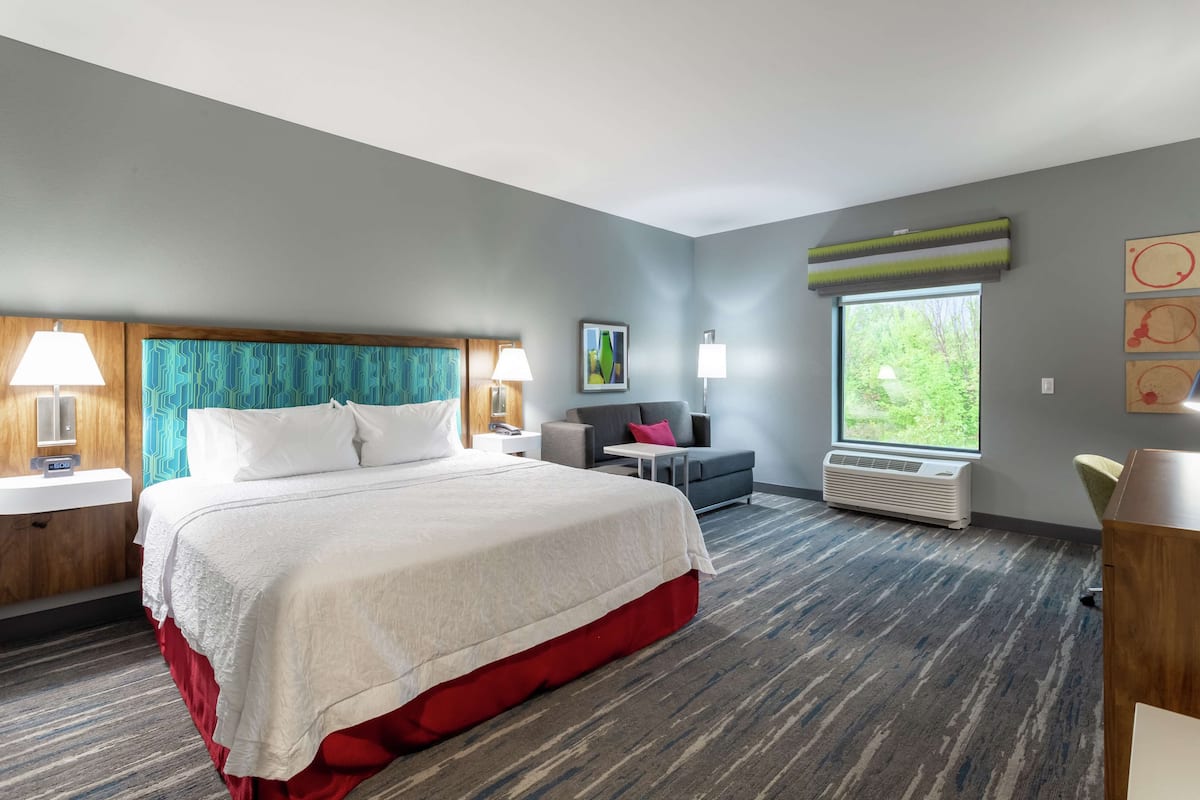 Une chambre d’hôtel avec un grand lit, un bureau, une chaise, une fenêtre et un climatiseur fixé au mur.