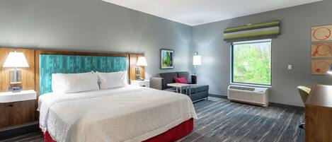 A hotel room with a large bed, a desk, a chair, a window, and a wall-mounted air conditioner.