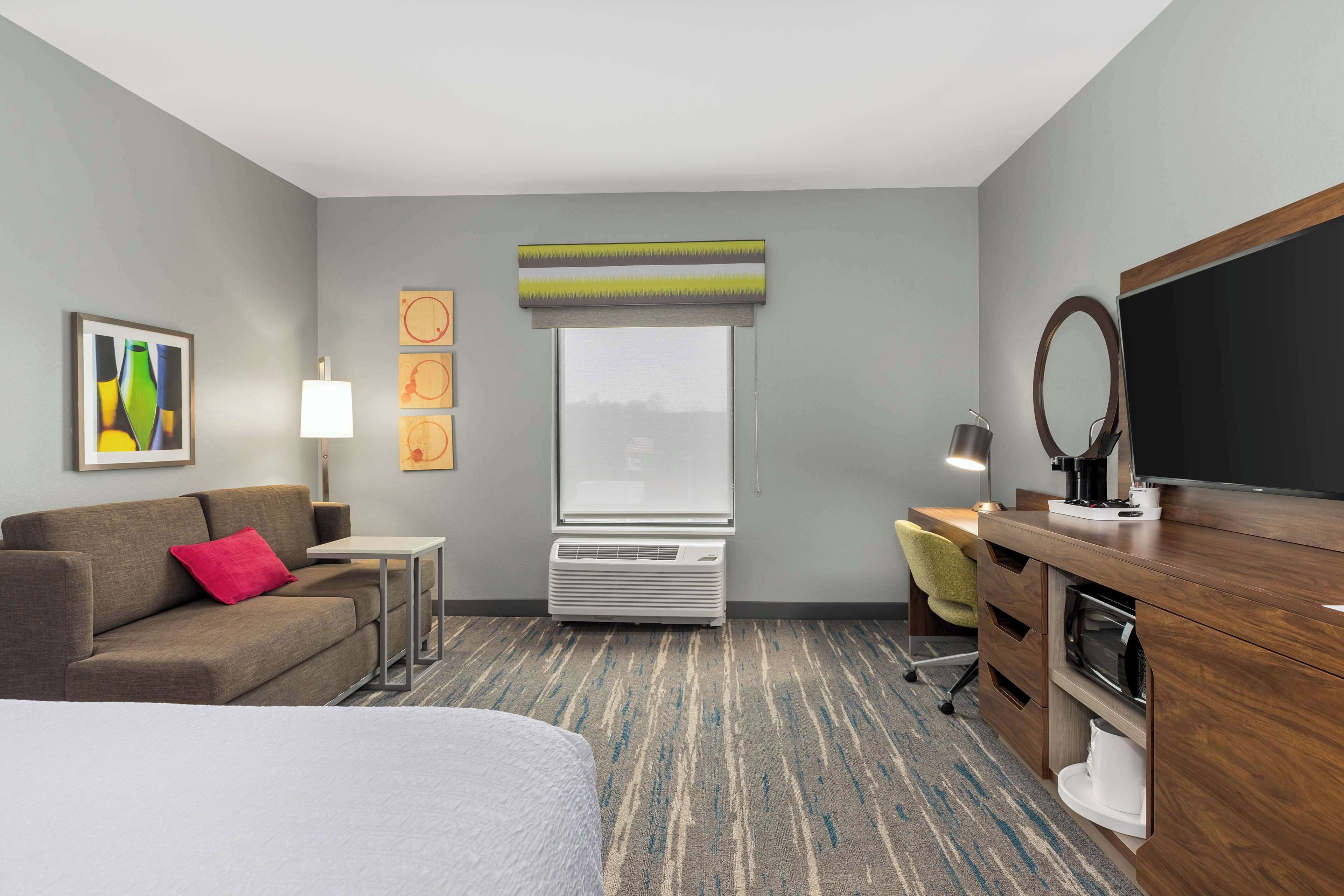 A hotel room with a bed, a sofa, a desk with a chair, a television, a window with blinds, and a wall-mounted air conditioner.
