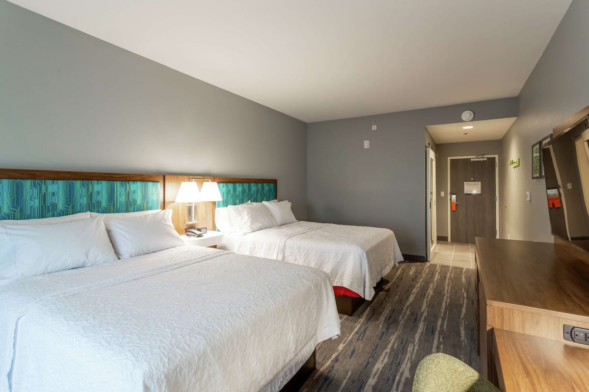 Une chambre d’hôtel avec deux lits, un bureau, une chaise et une salle de bain visible à travers une porte ouverte.