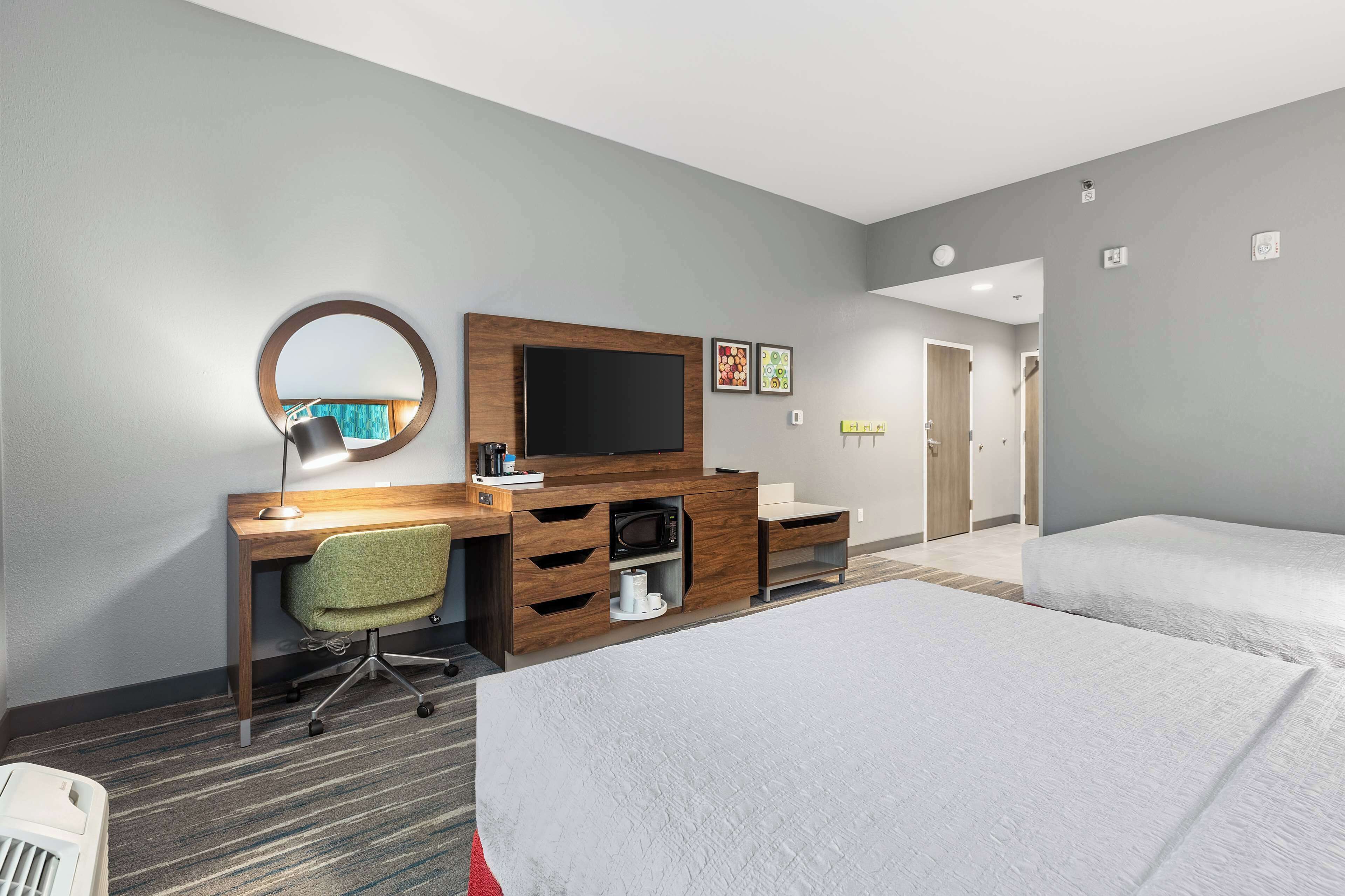 A hotel room with a wooden desk, a flat-screen TV, a green office chair, a microwave, a small cabinet, and two single beds.