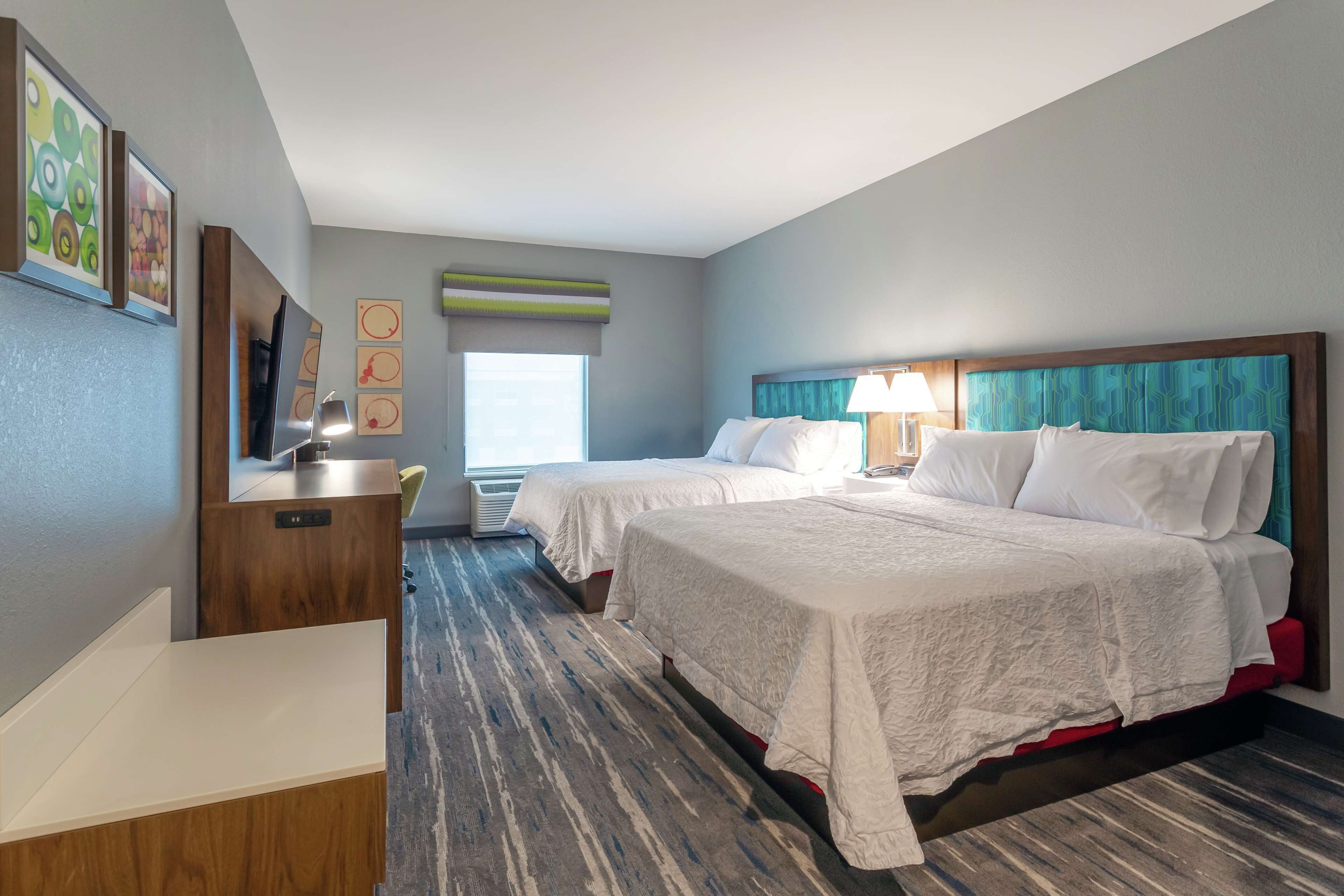 A hotel room with two beds, a desk, and a TV. There is a window with blinds and a view of the outside.