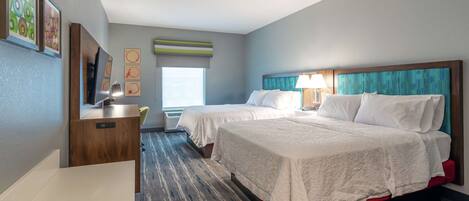A hotel room with two beds, a desk, and a TV. There is a window with blinds and a view of the outside.