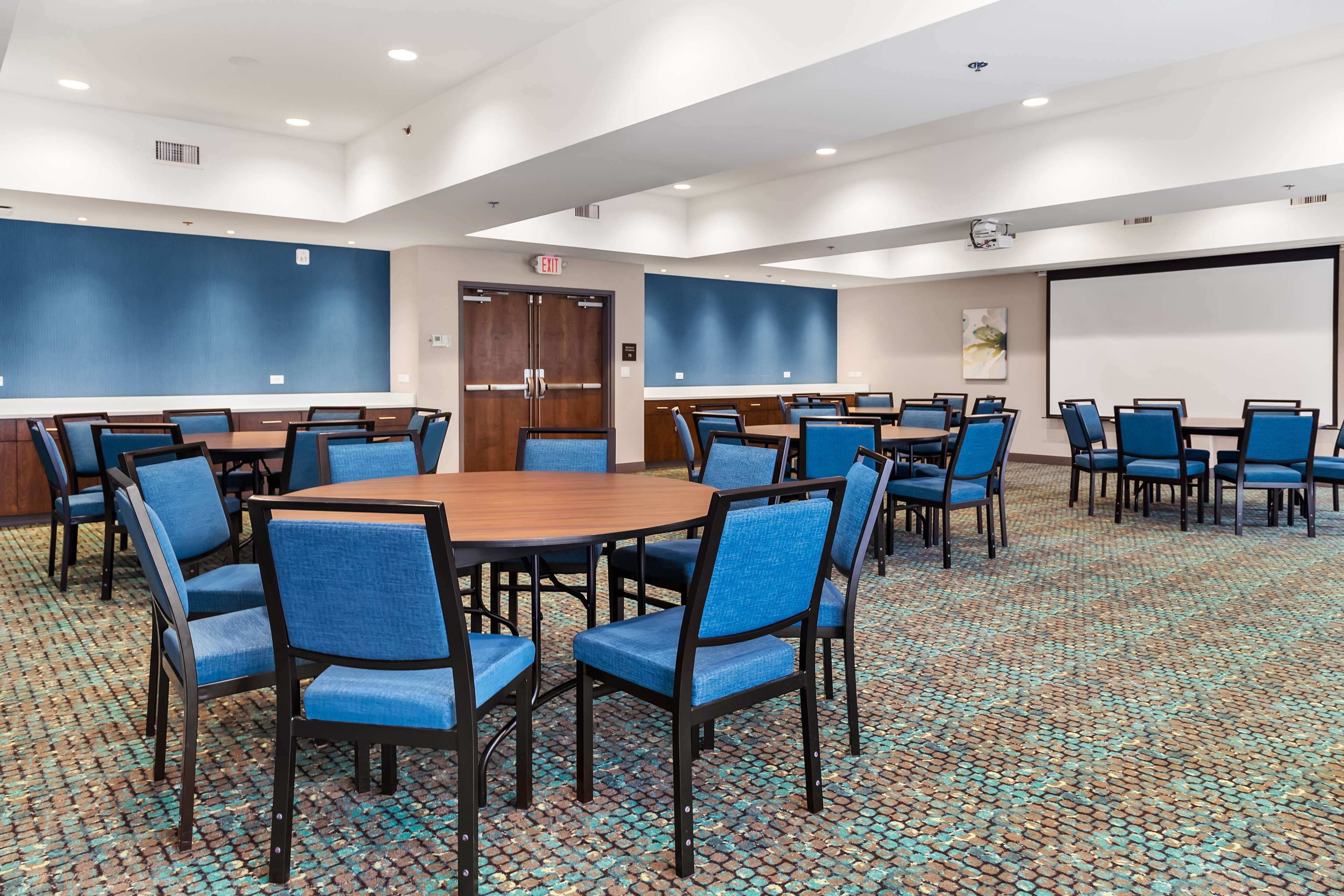 A conference room with round tables and blue chairs, a projector screen, and a large door.