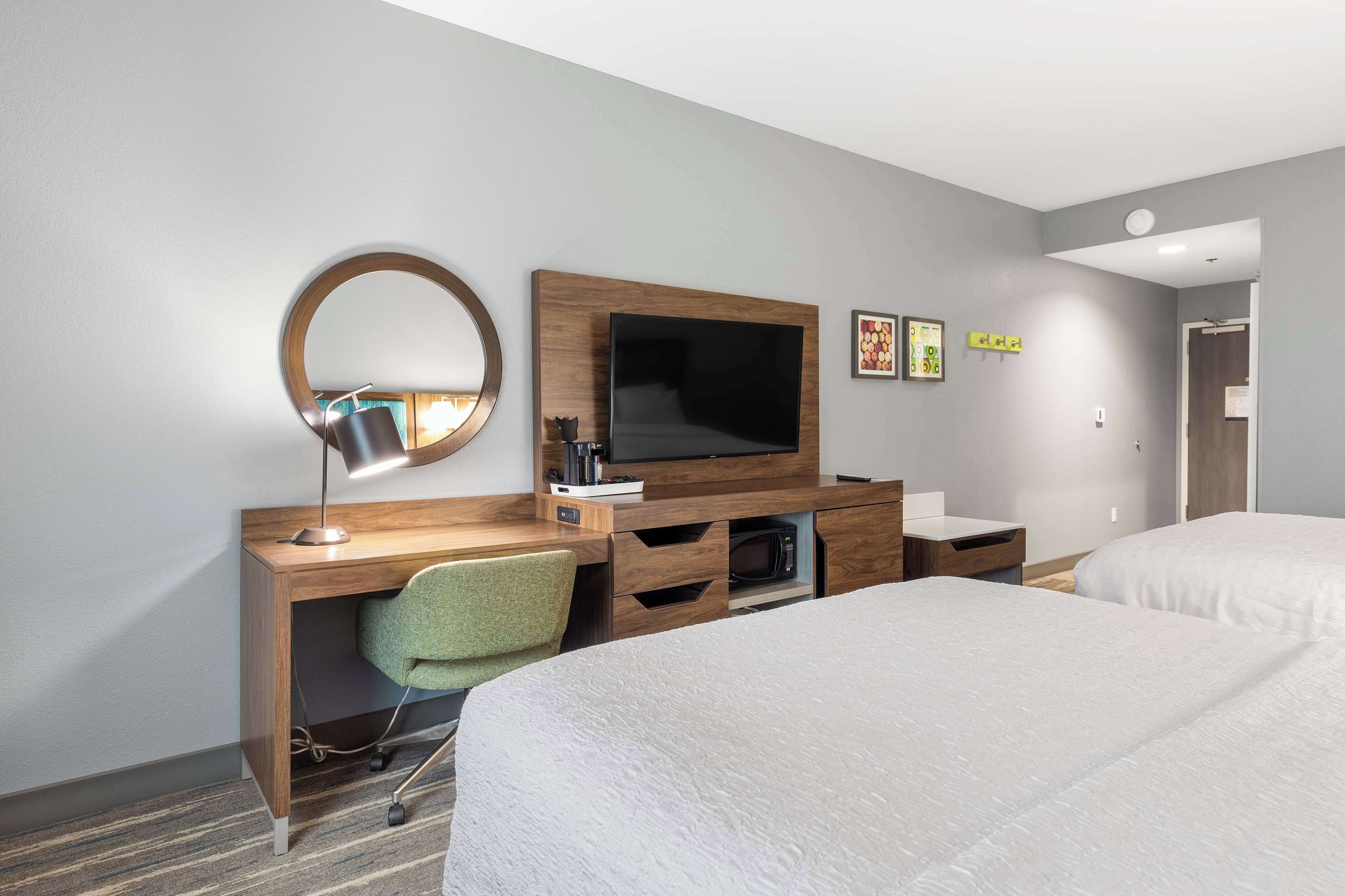 Habitación de hotel con un escritorio de madera, un espejo redondo, televisor de pantalla plana, una silla verde, dos camas y un baño pequeño.