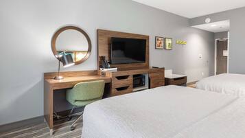 Habitación de hotel con un escritorio de madera, un espejo redondo, televisor de pantalla plana, una silla verde, dos camas y un baño pequeño.