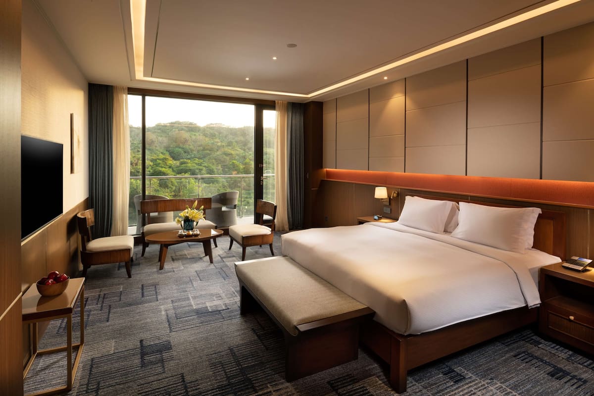 A hotel room with a large bed, a desk with a chair, a TV, and a view of a forested area.