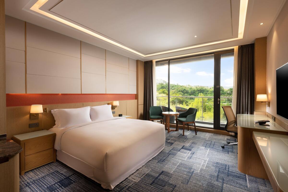 A hotel room with a large bed, a desk with a chair, a small table with two chairs, a TV, and a balcony with a view of greenery.
