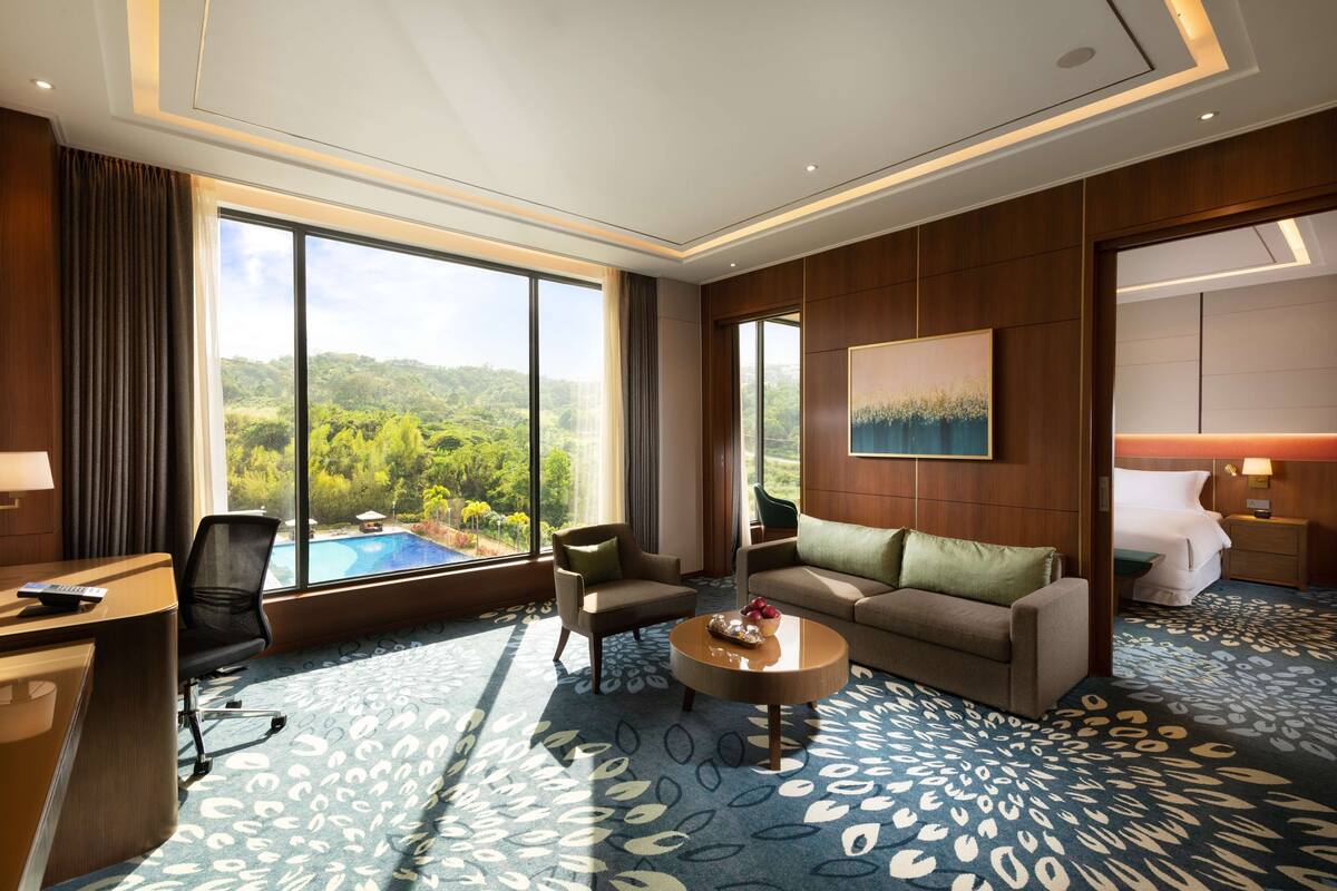 A hotel room with a large window offering a view of a pool and greenery, a sofa, a coffee table, and a desk with a chair.
