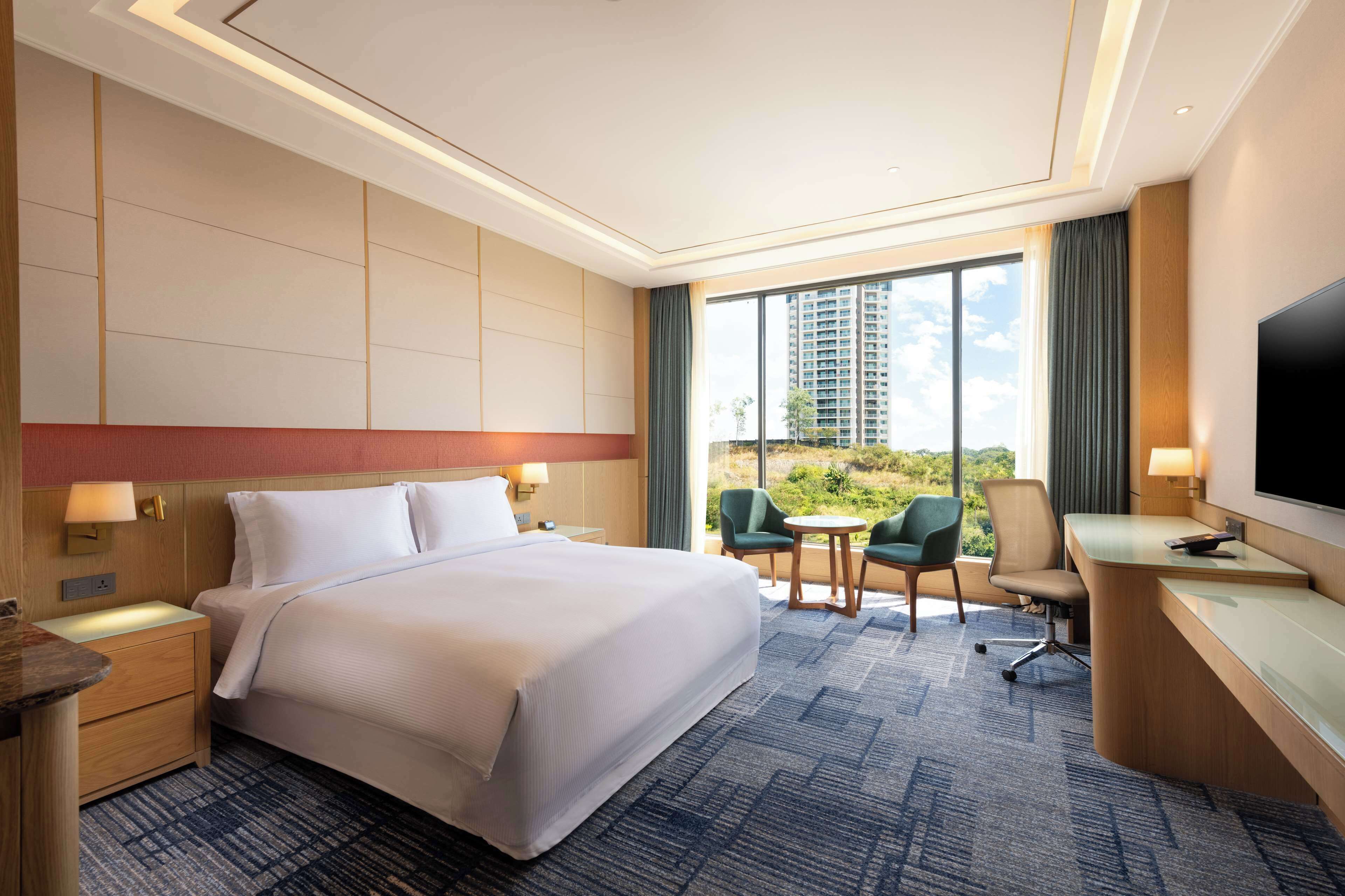 A hotel room with a large bed, a desk, two chairs, a TV, and a view of a cityscape through large windows.