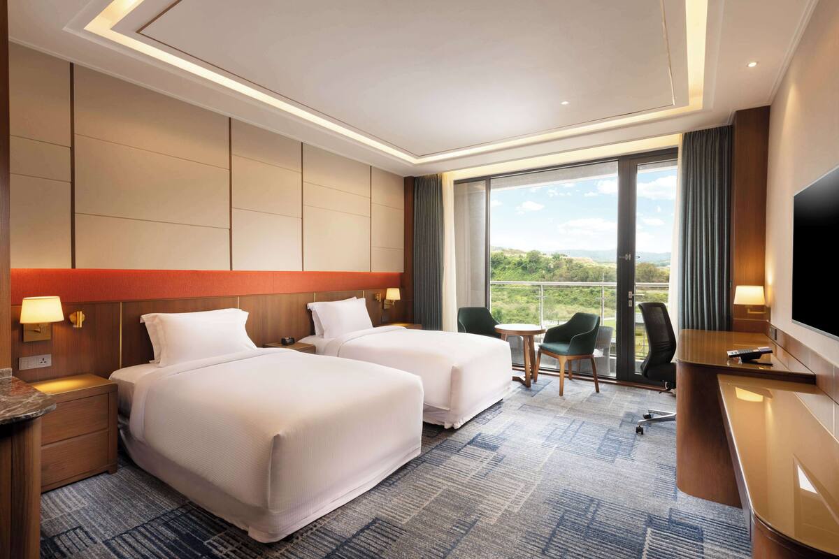 A hotel room with two beds, a desk, a TV, and a view of the outdoors.