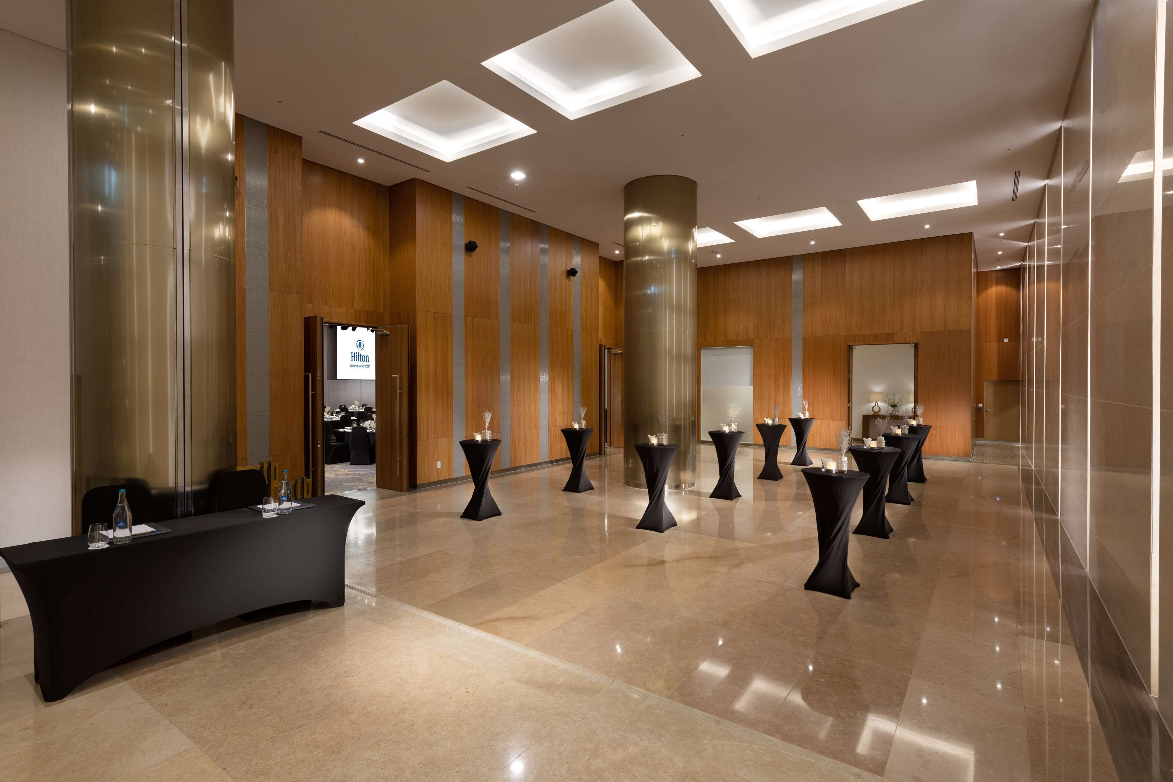 Um hall de hotel moderno com um balcão de recepção comprido, várias mesas redondas pequenas com velas e um piso de mármore polido.