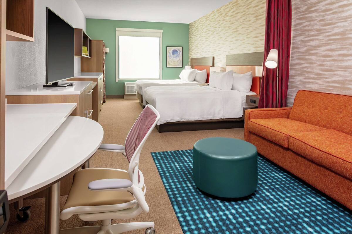A hotel room with a desk, two beds, a sofa, a chair, and a round ottoman.