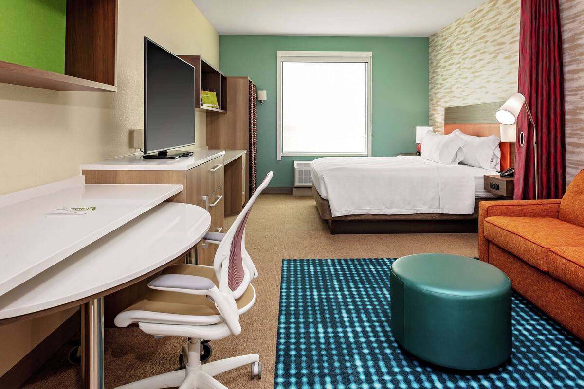 A hotel room with a bed, a desk with a chair, a television, a window, and a sofa.