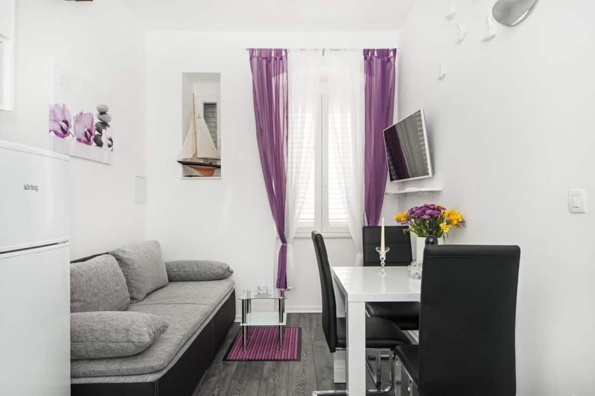 A compact living space with a sofa, a dining table with chairs, a TV, and a small window with purple curtains.