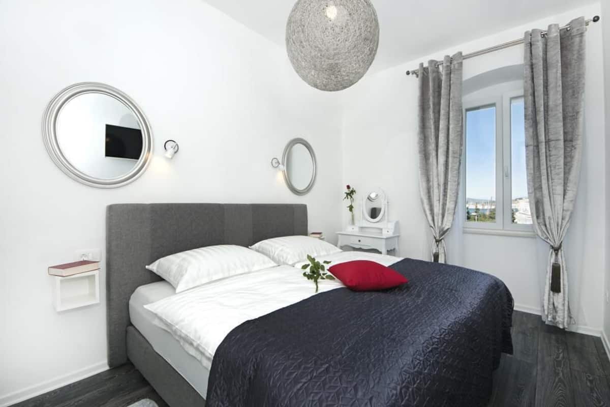 A modern bedroom with a grey headboard, a round mirror, a bedside table with a lamp, and a window with curtains.