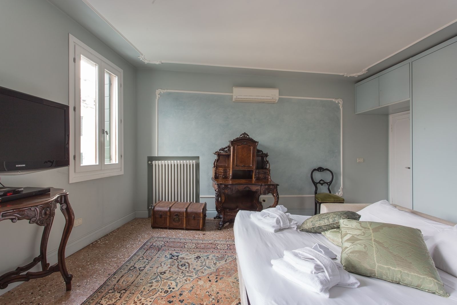 A room with a bed, a radiator, a wooden cabinet, a television, and a window.