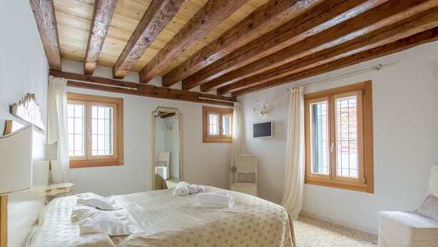 Ein Schlafzimmer mit einem großen Bett, Holzbalken, zwei Fenstern, einem Spiegel und einem Fernseher.