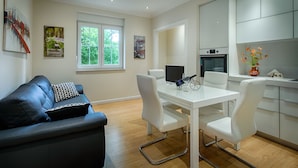 A modern living room with a black leather sofa, a white dining table with chairs, and a kitchen area with grey cabinets and a built-in oven. - Apartment Marella (Split)