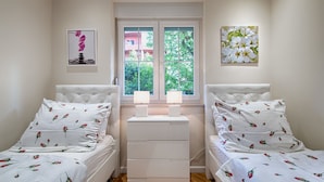 A bedroom with two beds, each with floral bedding, a bedside table with lamps, and a window with a view of greenery. - Apartment Marella (Split)