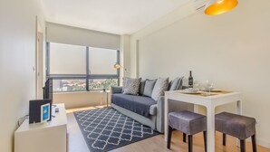 A modern living room with a sofa, a dining table with benches, a TV, and a large window offering a city view. - Trindade Flat with Great View (Porto)