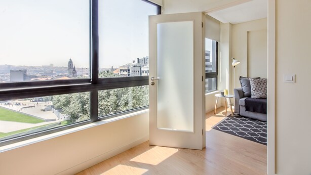A modern apartment with a large window, a door leading to a living area with a sofa and a lamp, and a view of a cityscape. - Trindade Flat with Great View (Porto)