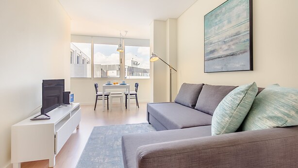 Un soggiorno moderno con un divano grigio, una TV a schermo piatto su una consolle bianca, un tavolo da pranzo con sedie e un'ampia finestra con vista sugli edifici.
