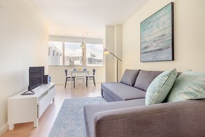 Un salon moderne avec un canapé gris, un téléviseur à écran plat posé sur une console blanche, une table à manger avec des chaises et une grande fenêtre donnant sur des immeubles.