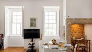 A dining area with a glass table, white chairs, a stone fireplace, and a television on a stand. - São Mamede Downtown Apartment (Lisbon)